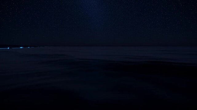 Glowing blue waves crash on a dark beach at night with stars shining brightly.