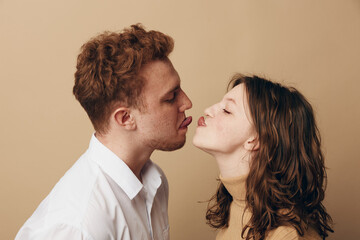 Young couple with curly hair playfully sticks out tongues facing each other on beige background....
