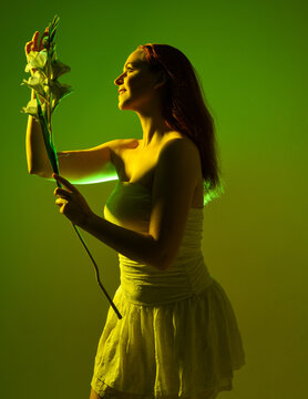Close up portrait of female model with red hair and white dress, holding flowers in side profile.  Isolated figure with colourful creative studio lighting with golden glowing silhouette and green back