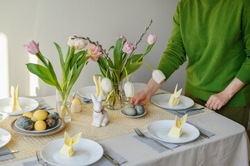 Woman in green sweater serving table for Easter holiday family dinner. Traditional Easter spring table setting with green, yellow and blue dyed eggs, rabbit figurines and flowers, home celebration
