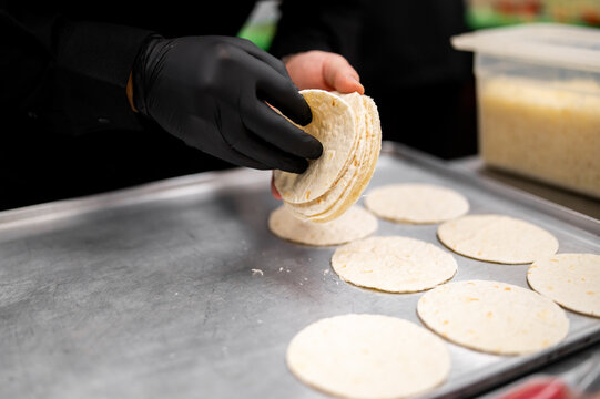 A person wearing black gloves is shown placing small white corn tortillas onto a metal baking sheet in a commercial kitchen setting.