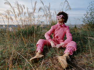Naklejka premium Outdoor person wearing pink fashion sits among tall grasses in a field, relaxed pose with boots, wide sky, natural setting, calm mood, candid lifestyle scene for travel or editorial use