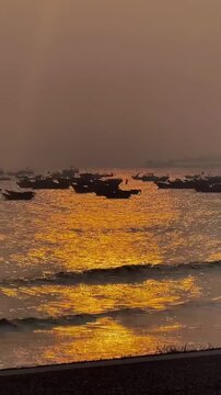 A stunning golden sunrise over the sea in Weihai, China. The ocean surface is bathed in orange and gold light with glittering ripples. Fishing boat silhouettes add a cinematic touch to this coastal mo