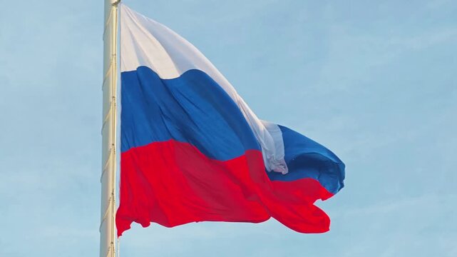 The Russian Federation flag billows dynamically in the wind on a tall flagpole, with the blue sky as a backdrop. National pride concept.