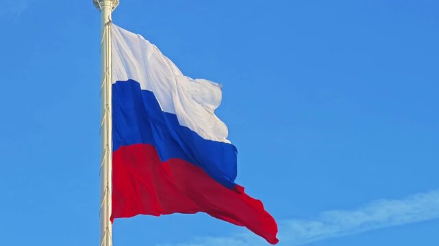 The Russian Federation flag, with its white, blue, and red stripes, flutters proudly on a flagpole against a beautiful blue sky. National symbol.