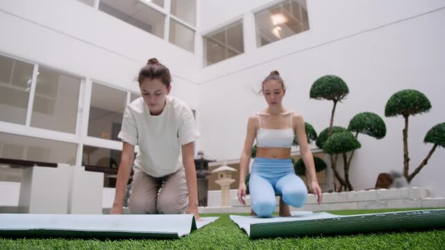 Two girls roll out yoga mats and sit in lotus pose in the inner courtyard, preparing for joint meditation.
