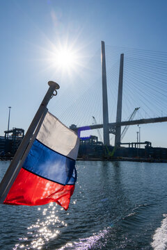 Russian flag waving in the foreground with the Zolotoy Bridge and sun shining over the water in Vladivostok, Russia