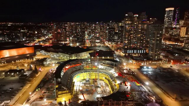 A rotating aerial night view of a sports arena under construction with the illuminated city skyline in Calgary, Alberta, Canada.