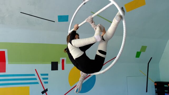 Nine year old girl practices aerial gymnastics tricks on aerial hoop in sports school with safety harness and elastic bandages on legs, slow motion, strength flexibility training