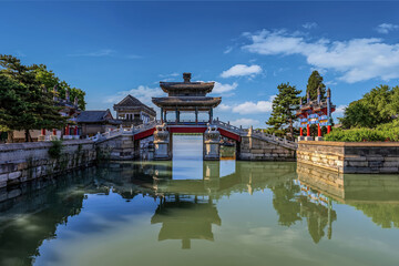 Summer view of a traditional arched bridge and pavilion reflected on calm water at the Summer Palace in Beijing, China under blue sky.