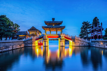 Illuminated Xingqiao Bridge at the Summer Palace in Beijing on a summer night, traditional Chinese architecture reflected in calm water.