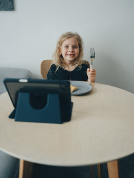 A child at the table eats breakfast and watches entertainment shows on a tablet.