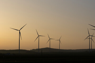 Naklejka premium a wide shot of several large wind turbines silhouetted against a hazy sunset sky over a dark rural field in a calm evening atmosphere