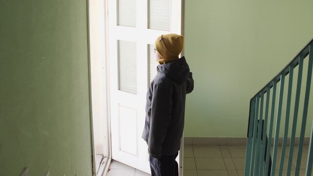 Young boy in winter hat and jacket opening an apartment door into a bright hallway, pausing at the threshold as he prepares to leave home for school or a new day