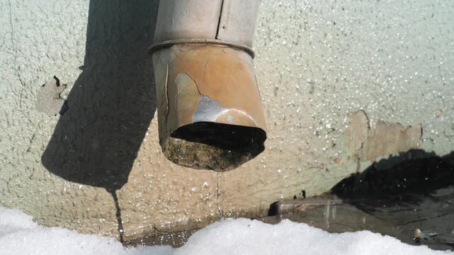 Rusty downspout corroding and shedding paint, dripping melting snow into bright sunlight, puddles form on concrete near icy patches during thawing winter day