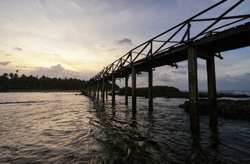 Fototapeta premium A wooden pier overlooks the calm ocean waves as the sun sets, creating a serene and tranquil tropical atmosphere, perfect for relaxation and contemplation with palm trees on the horizon.