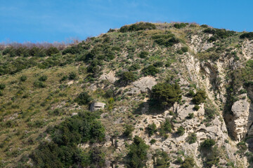 Scenic mountain slope with green vegetation, rocky cliffs, and an old bunker under a clear blue sky