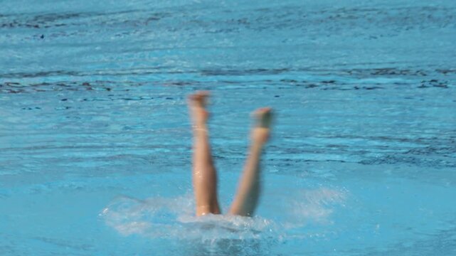 Artistic swimmer performing choreographed leg movements upside down in clear blue swimming pool, synchronized swimming training underwater sport practice routine