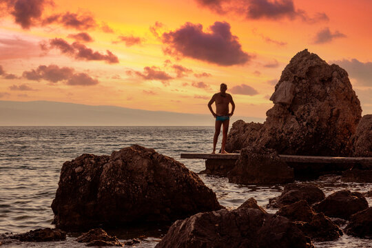 A male swimmer silhouetted against the evening sky as he gazes out across the sea