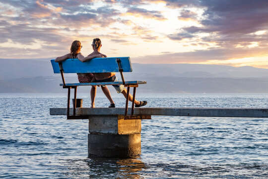 A seated couple enjoy the view of the sea as the sun sets