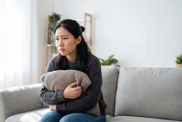 Obraz premium Sad asian woman hugging a pillow while sitting alone on a sofa