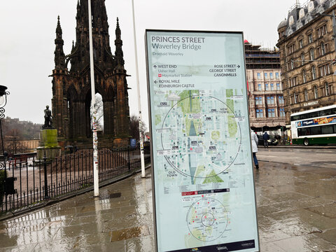 Tourist map kiosk and Scott Monument on Princes Street Waverley Bridge in Edinburgh, Scotland, UK on a rainy overcast day with wet pavement and city bus