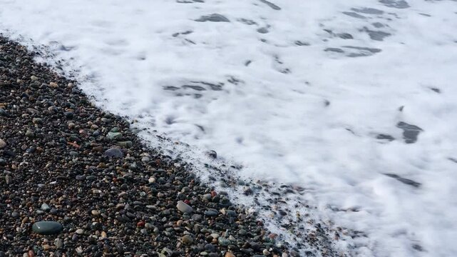 A serene, horizontal shot of a gentle turquoise wave breaking into soft white foam on a dark pebble beach. The clear water of the Black Sea stretches toward the horizon under a soft, diffused light