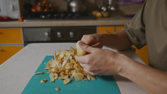 Close view of hands removing potato skin with a vegetable peeler above a cutting board. Thin strips of peel fall onto the board during the peeling process. High quality 4k footage