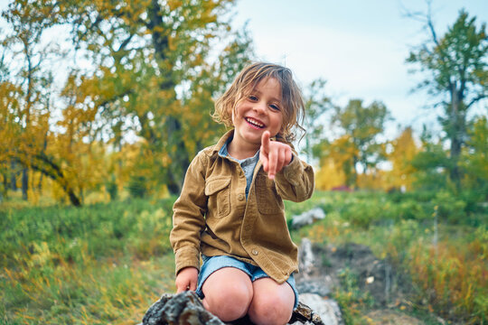 Young boy sitting on a log, pointing at the camera with a smile on his face.