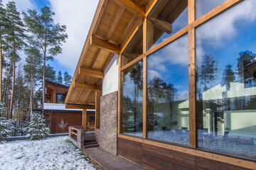 Fototapeta premium A building made of wood and stone sits among tall trees in a forest area. Snow covers the ground while blue skies are visible through large windows of the structure