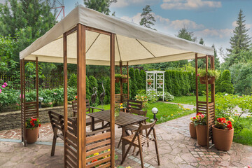 A wooden shelter with a white canopy stands in a garden filled with plants and flowers. Wooden chairs and a table occupy the space. The sky appears cloudy and overcast © Alhim