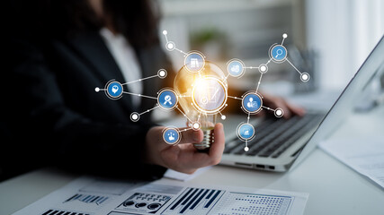 Business professional holding a glowing light bulb with digital icons representing innovation,...