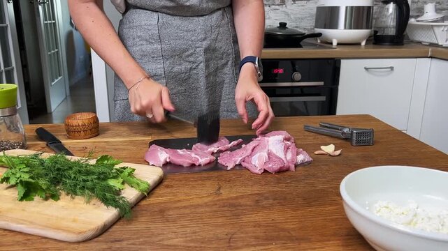 Hands using a black spiked meat hammer to tenderize raw pork slices on a metal board, kitchen preparation scene