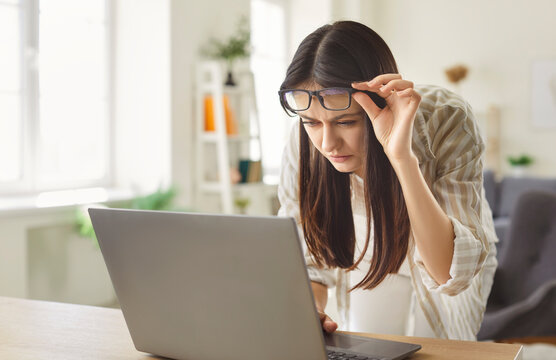 Young woman leaning toward laptop and lifting glasses while struggling to read text. Focused female with myopia struggling with blurry vision, experiencing eye strain and discomfort while working.