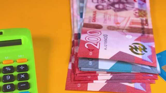 A stack of 200 Ghana Cedi banknotes drops sits on a yellow background alongside a green calculator and a credit card. This clean, top-down shot represents banking, savings, and West African commerce.