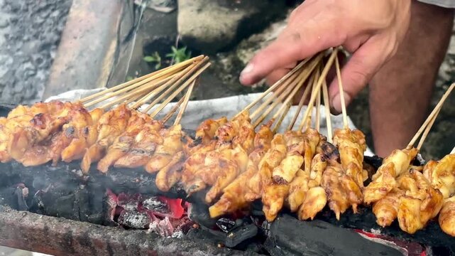Chicken satay skewers grilling over a traditional street food charcoal grill. Hands cooking Indonesian satay on a roadside food stall.