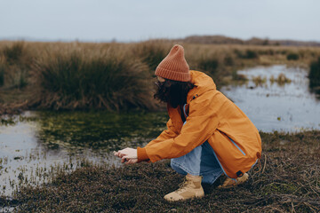 Woman lifestyle concept showing a woman in an orange jacket and beanie crouching by a wetland,...