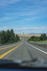 Clear sky above endless highway stretches ahead. Yellow lines divide lanes, guiding steady motion. Trees flank sides, green against golden fields. Distant hills fade softly under broad blue