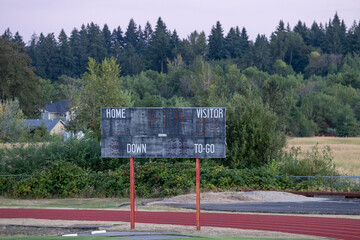 Old scoreboard stands idle in quiet field. Red track curves beside it, empty and still. Trees form a green backdrop, dense and tall. House sits nestled behind bushes, small and quiet