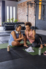 Diverse female friends sitting on green mats at gym wearing workout clothes checking smartphone