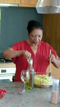 Vertical video: Stirring red-blouse cook mixing pasta kitchen bowl as green helper entering to help