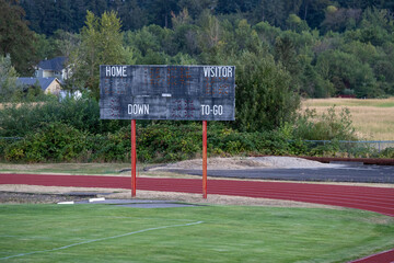 Empty scoreboard stands beside a grassy field. Red track surrounds the playing area. Trees and houses dot the background. No people are visible. Ideal for sports, recreation, or quiet park themes
