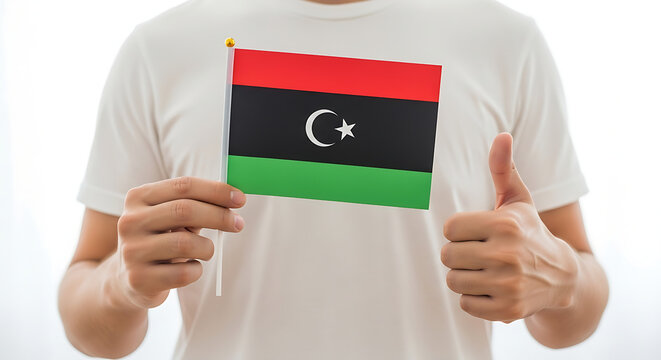 ​A person in a white shirt holding a small Libyan flag while giving a thumbs up against a bright background.