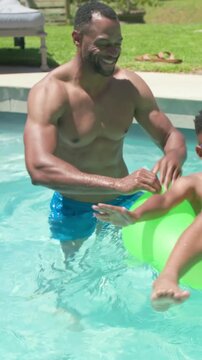 Vertical video: Entering backyard pool, dad in trunks guiding child in green ring safely, goggles