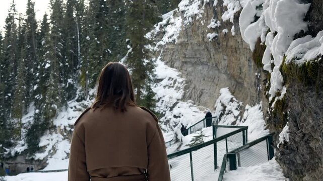 A young woman walks along a path in a snowy mountain forest. She pauses to look around, appreciating the natural winter beauty. Perfect for travel, adventure, or lifestyle projects. Johnston canyon