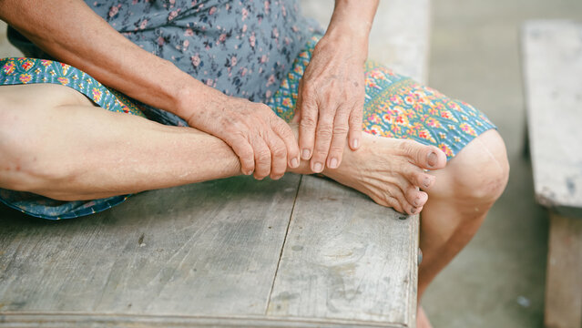 Close up of senior woman massaging painful ankle while sitting on wooden bench. Elderly foot with joint pain, inflammation and swelling. Healthcare, podiatry and chronic arthritis concept.