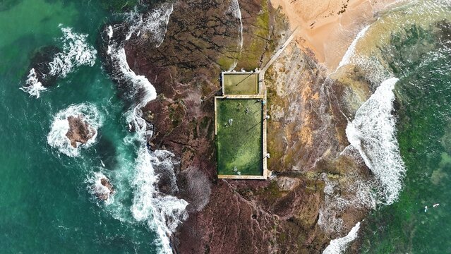 Drone shot aerial view of a beach rock pool and waves crashing over rocks at a beach in Sydney Australia in Summer 