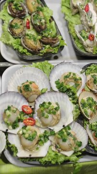 Vertical screen Fresh Abalone And Scallops On Lettuce Trays, Chef Grills And Seasons With Garlic Butter And Chopped Green Onion, Oyster Shells Glinting, Marketplace Aroma And Gourmet Street