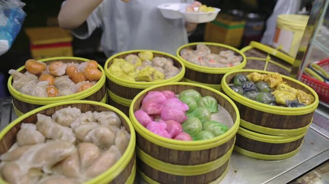 Busy Vendor Serving Dim Sum At Street Market, Closeup Of Hands Using Tongs To Transfer Steamed Dumplings Into Takeaway Box, Rows Of Bamboo Steamers With Colorful Buns, Lively Evening Stall