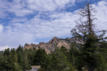 Obraz premium Rugged mountain peak rises sharply against blue sky. Rocky slopes host scattered pine trees and wild shrubs. Wilderness feels untouched, untamed, and serene. Crater Lake National Park in Oregon, USA.